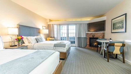 A bright hotel room with two double beds, a fireplace, a work desk, and a large window with a balcony view, all in soft neutral tones.