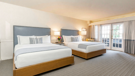 Two neatly made hotel beds with gray headboards, nightstands, and lamps, plus a bright room with a large windowed door to a balcony.