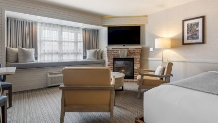 A cozy hotel suite with a brick fireplace, comfortable seating, a TV above the fireplace, a window seat, and a neatly made bed ending the sentence.