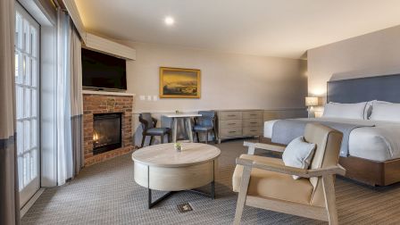 Modern hotel suite with two queen beds, a seating area, fireplace, flat-screen TV, and large balcony doors letting in natural light.