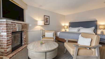 A cozy hotel room with a brick fireplace, a mounted TV, a king bed, two beige armchairs, a round coffee table, and soft lighting, creating a warm, inviting space.