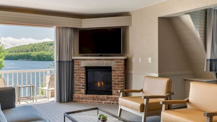 A modern living room with a stone fireplace, mounted TV, two tan armchairs, a glass coffee table, and a view of a lake through large glass doors.