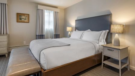 A neatly made hotel room with a large bed, nightstands with lamps, a bench at the foot, and soft, neutral d&eacute;cor.