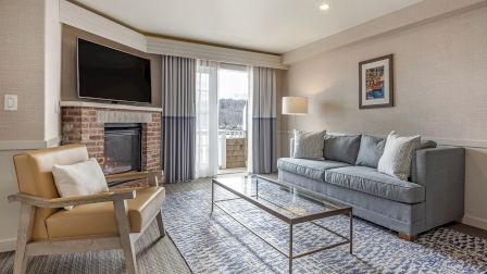 A modern living room with a gray sofa, beige armchair, glass coffee table, brick fireplace with a mounted TV, and a sliding glass door opening to a balcony.