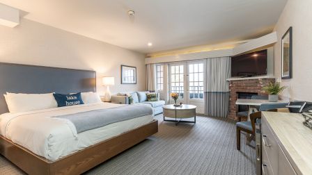 A spacious hotel room with a king bed, seating area, desk, flat-screen TV, Sunday-style decor, and a fireplace/TV nook by the windows, ending.