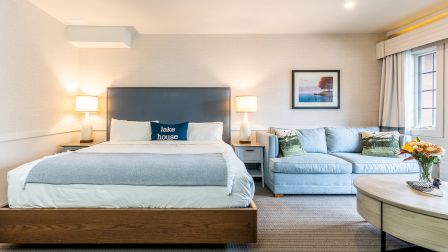 A modern hotel room with a king bed, two nightstands, a blue-gray headboard, and a light gray sofa with throw pillows; a coffee table and vase of flowers.