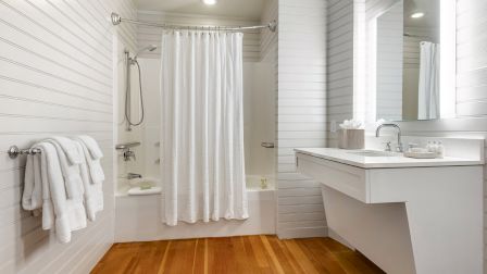 A bright bathroom with white paneled walls, a white shower curtain, a tub with a handheld shower, a towel rack with fluffy towels, and a modern white vanity with a mirror, all on warm wooden flooring.