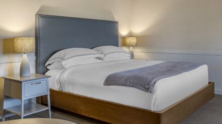 A tidy hotel bedroom with a large wooden bed, gray headboard, white linens, a folded blanket, and two bedside lamps on matching nightstands.