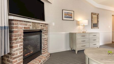 Cozy living room with a brick fireplace, mounted TV, light gray furniture, dresser with lamp, framed art, and a round coffee table.