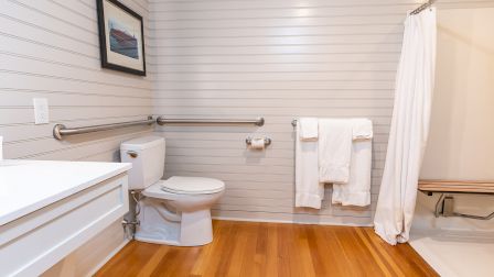 A clean bathroom with a white toilet, grab bars, a wooden floor, a white sink, towel rack, a shower with a curtain, and a bench in the tub area.