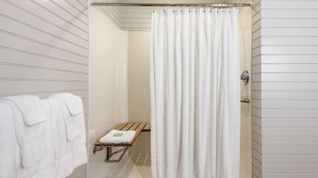 A bright shower stall with a white curtain, striped walls, and a small bench with folded towels inside the cubicle.