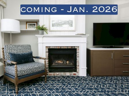 A cozy room with a chair, lamp, fireplace, and TV, featuring a "Coming - Jan. 2026" sign above.