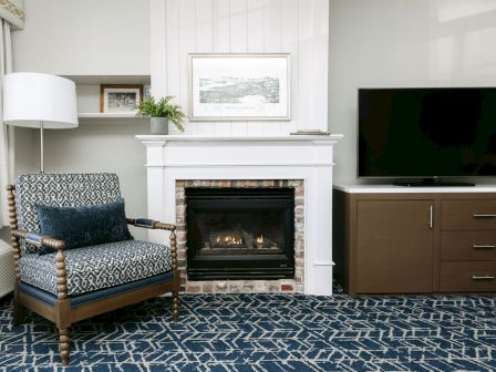 A cozy living room with a fireplace, a patterned blue rug, a patterned armchair, a tall floor lamp, a framed artwork above the mantel, and a TV on a brown cabinet.