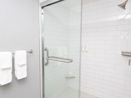 A clean, glass-walled bathroom shower with white subway tiles, a chrome grab bar, a shower head, and two white towels on a rack outside the glass door.