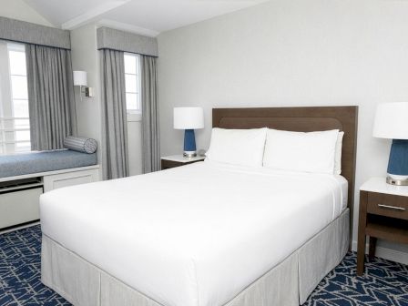 A clean hotel room with a white bed, two bedside tables and lamps, a wooden headboard, and a windowed seating area with blue accents.