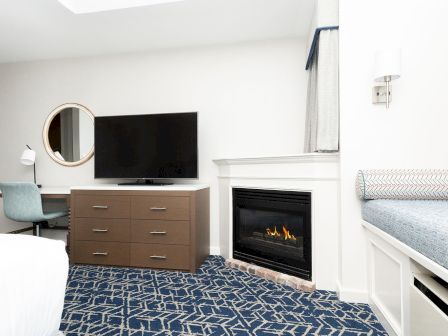 A bright hotel room with a TV above a brown dresser, a round mirror, a desk and chair, a fireplace, and a blue patterned carpet.