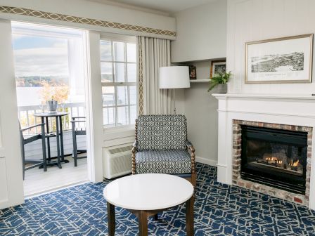 A cozy hotel living area with a fireplace, patterned blue carpet, chair, round white table, and a balcony view through glass doors.