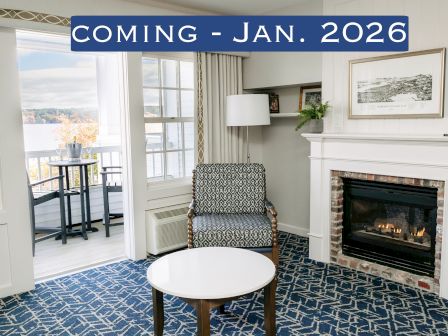 A cozy room with a fireplace, armchair, and table. Balcony view outside. Text reads "COMING - JAN. 2026."
