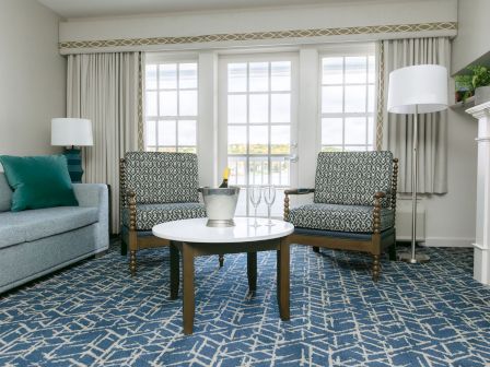 A cozy living room with a blue geometric carpet, gray sofa, two patterned armchairs, a round coffee table, and a fireplace with a lamp and plants.