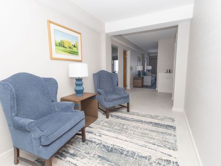 Cozy hotel-style hallway with two blue armchairs, a side table and lamp, framed landscape, and a patterned rug along a light, airy corridor.