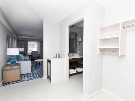 A bright hotel-like room with a living area, blue accents, a small desk, and an open bathroom; minimalist, airy, and modern.