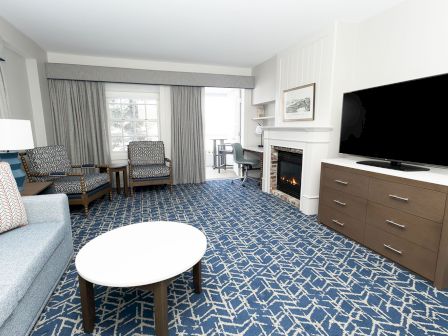 A bright hotel living room with a blue geometric carpet, sofa, two chairs, a round coffee table, and a large TV on a dresser.