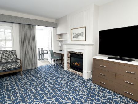 A cozy hotel suite with a blue patterned carpet, seating by a window, a fireplace, a desk, and a large flat-screen TV on a dresser.
