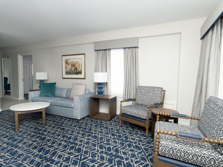 A tidy hotel living room with a blue patterned carpet, a light-blue sofa, two patterned armchairs, two lamps, a coffee table, and large windows with sheer curtains.