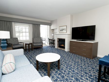 A cozy hotel living room with a blue patterned carpet, gray sofa, armchairs, round coffee table, and a fireplace with a flat-screen TV on a dresser.