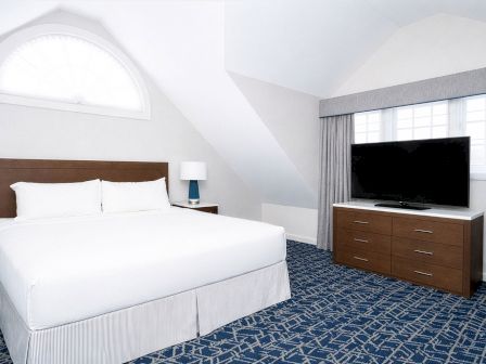 A bright hotel room with a queen bed, nightstand, table lamp, dresser, and a flat-screen TV on a dresser across from the bed.