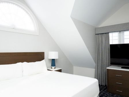 A bright hotel room with a white bed, wooden headboard, nightstand with a lamp, slanted ceiling, a large window arch, and a dresser with a flat-screen TV, all in a clean, modern style.