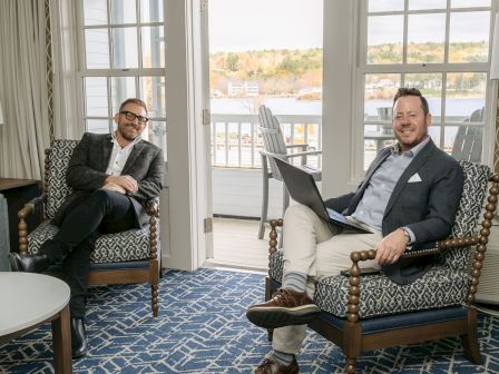 Two men in a bright hotel lounge, dressed in suits, sit in patterned chairs with a laptop and a relaxed vibe, chatting or posing.