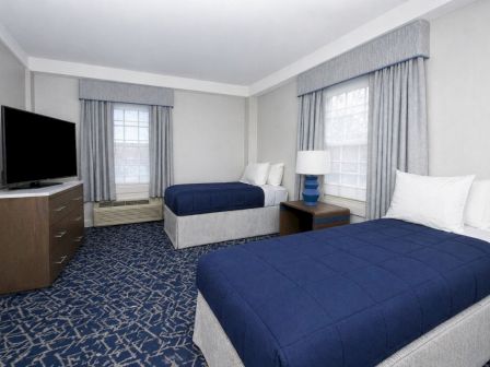 Two double beds with blue comforters in a hotel room, nightstand, lamp, dresser, and windows with blue curtains.