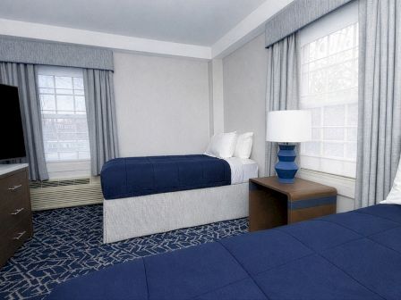 A hotel room with two double beds, blue bedspreads, white pillows, a nightstand with a blue lamp, a dresser, and windows with blue curtains.