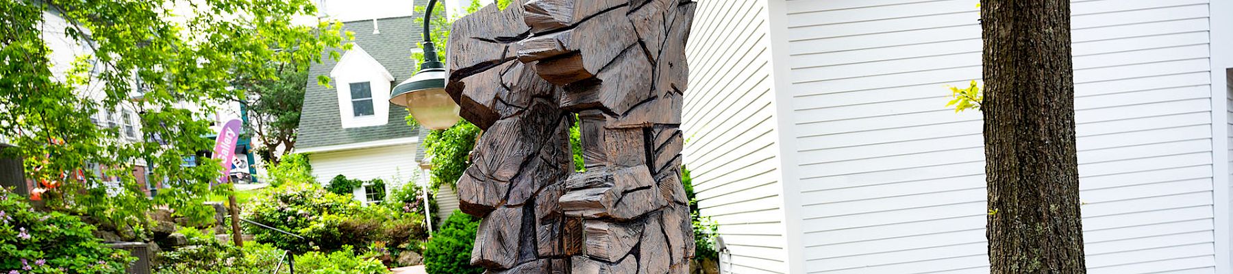 A weathered wooden sculpture of a cowboy-like figure stands in a landscaped garden beside a white building and steps.