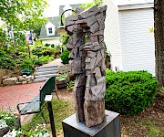 A weathered wooden sculpture of a cowboy-like figure stands in a landscaped garden beside a white building and steps.