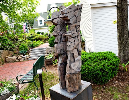 A weathered wooden sculpture of a cowboy-like figure stands in a landscaped garden beside a white building and steps.