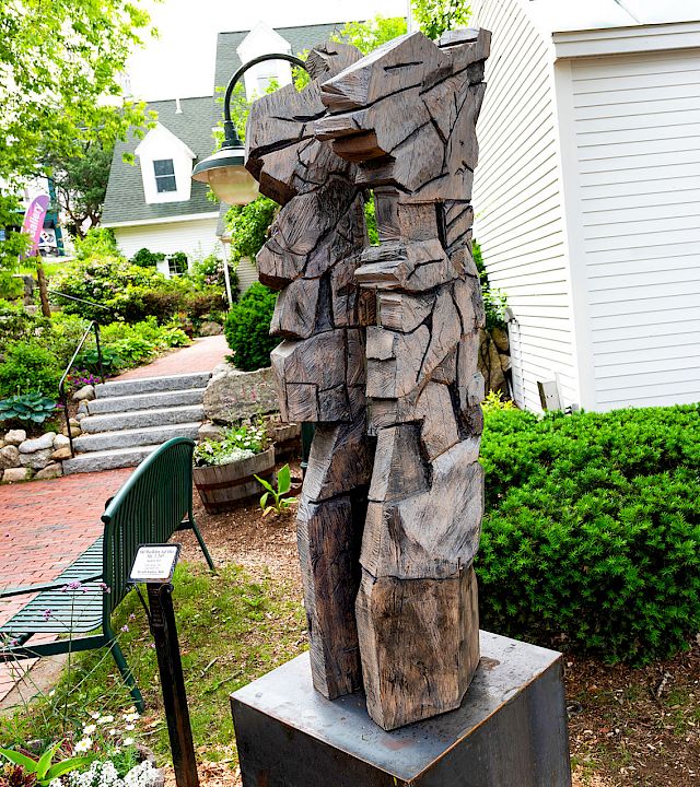 A weathered wooden sculpture of a cowboy-like figure stands in a landscaped garden beside a white building and steps.