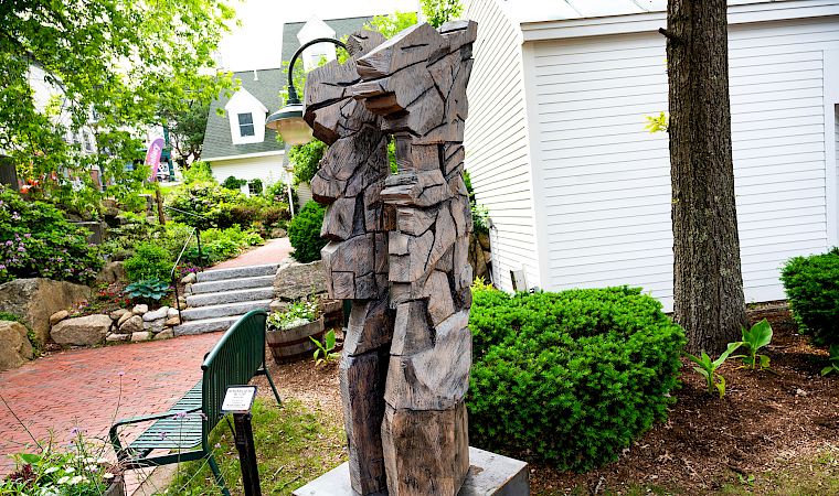 A weathered wooden sculpture of a cowboy-like figure stands in a landscaped garden beside a white building and steps.