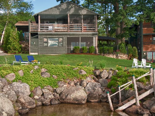 A lakeside house with a large porch, lawn with blue lounge chairs, white chairs, rocks, and a wooden dock beside the clear water's edge.