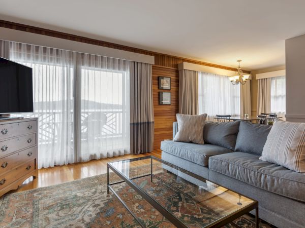 A cozy living room with a gray couch, glass coffee table, flat-screen TV, and large windows with sheer curtains, connected to a dining area.