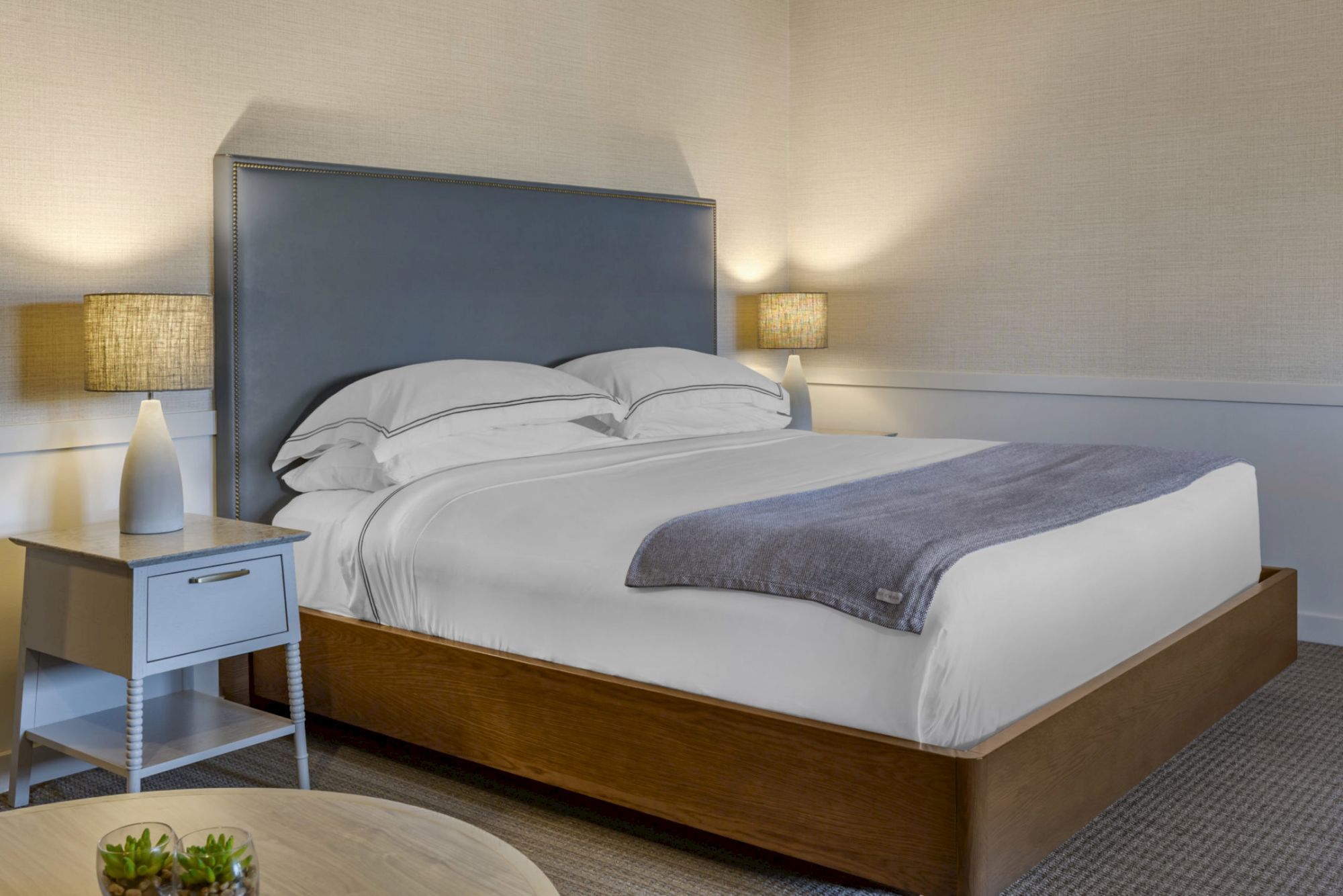 A cozy bedroom features a neatly made bed with a gray headboard, flanked by two lamps on nightstands. A small plant is on a nearby table.