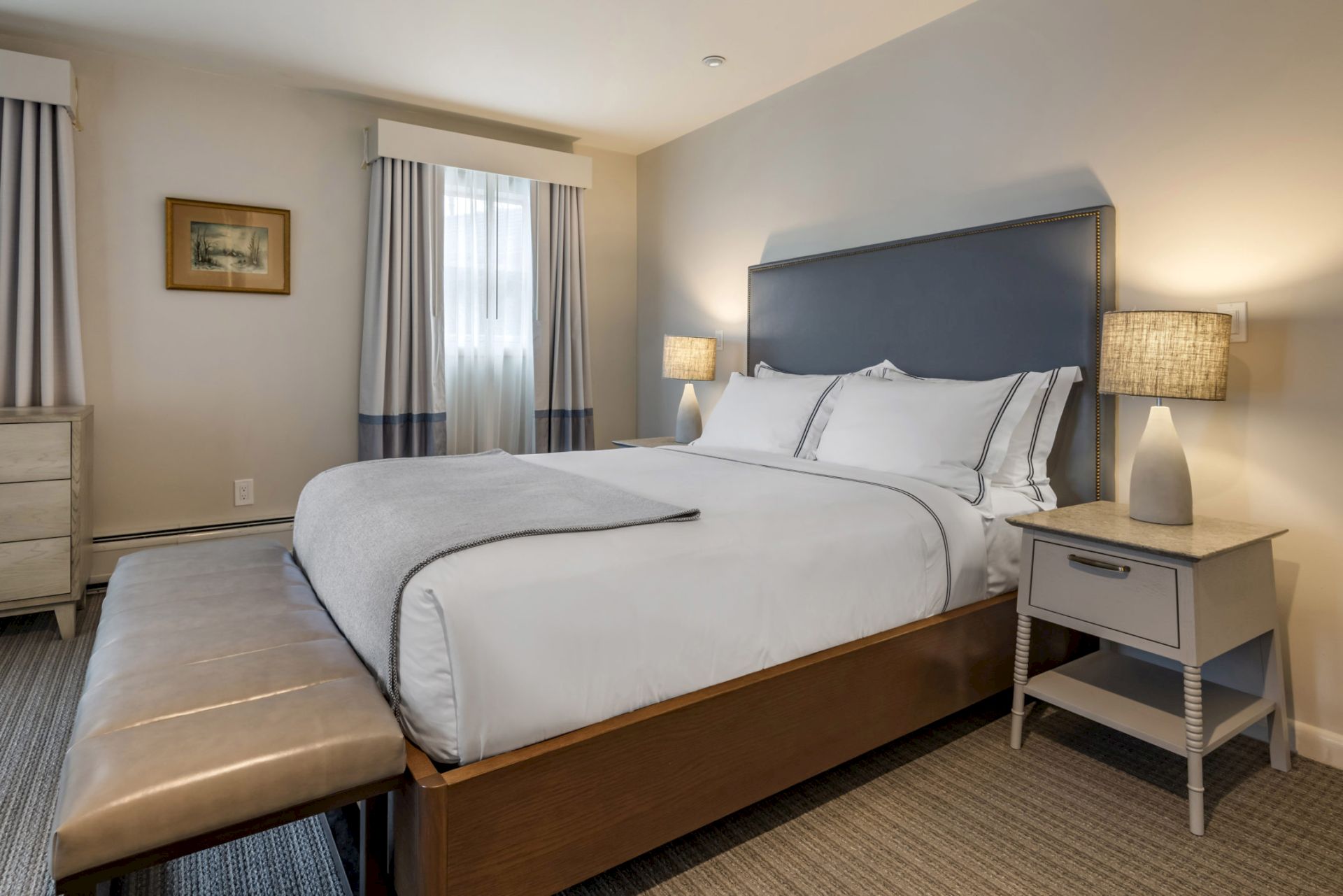 A neatly arranged bedroom features a large bed with white linens, bedside tables with lamps, a bench at the foot, curtains, and a framed picture.