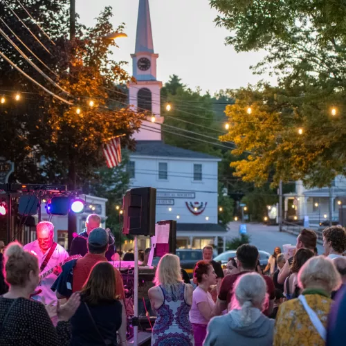 Outdoor concert at dusk with a band on stage, colorful lights, and a crowd watching in front of a clock-tower church.