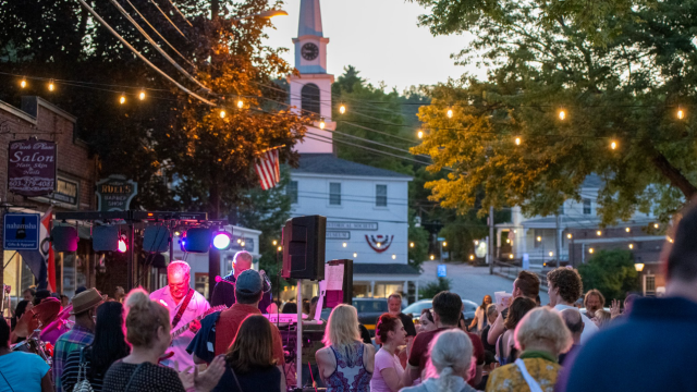 Outdoor concert at dusk with a band on stage, colorful lights, and a crowd watching in front of a clock-tower church.