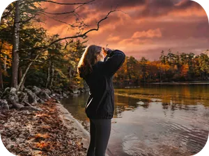 A person in a coat takes a photo of a calm lake at sunset, with autumn trees and cloudy skies all around.