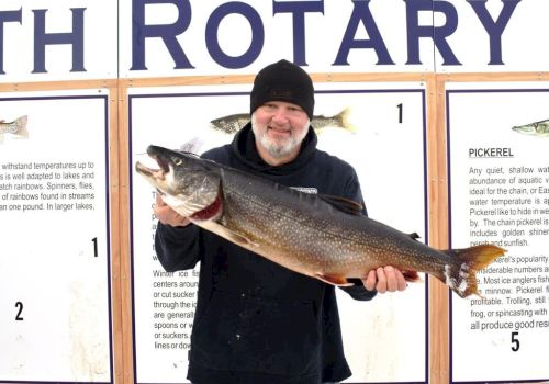 An older man in a black beanie holds a large fish in front of a &ldquo;FISH ROTARY&rdquo; display at a contest or event ending.