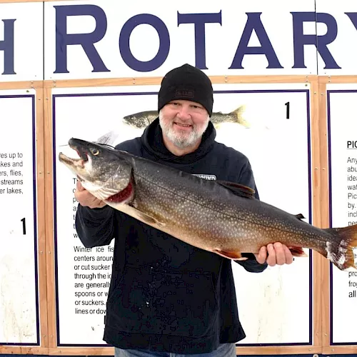 An older man in a black beanie holds a large fish in front of a &ldquo;FISH ROTARY&rdquo; display at a contest or event ending.
