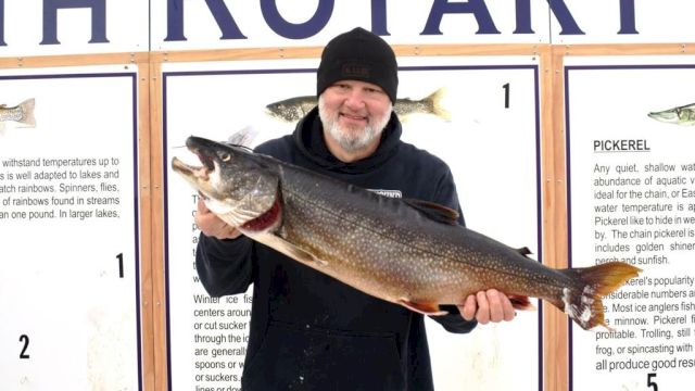 An older man in a black beanie holds a large fish in front of a &ldquo;FISH ROTARY&rdquo; display at a contest or event ending.