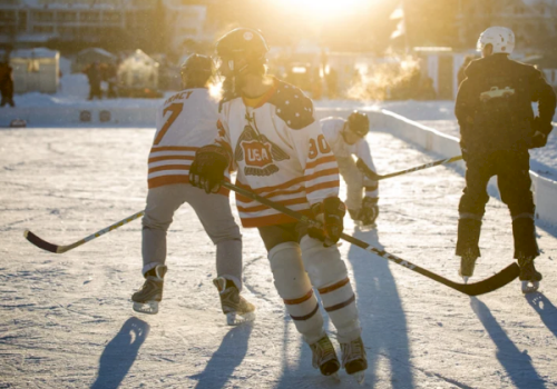 A hockey player in Team USA gear skates on an outdoor ice rink, with teammates and shadows stretching in bright sunlight, too.